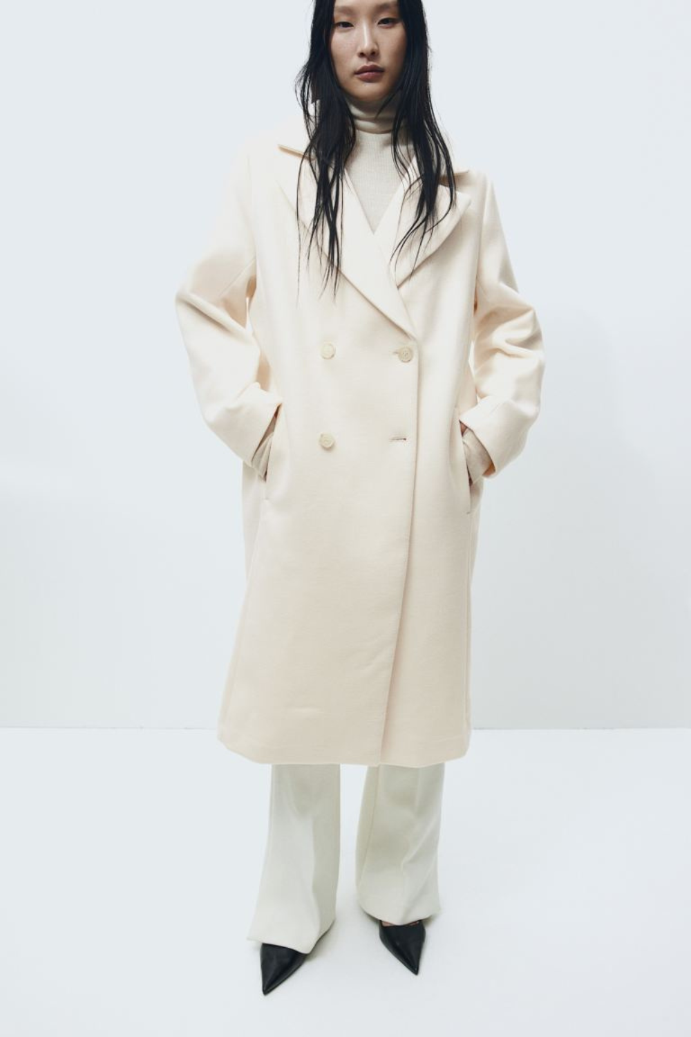 Model wearing a long cream overcoat with hands in pockets, white pants, and black pointed shoes.
