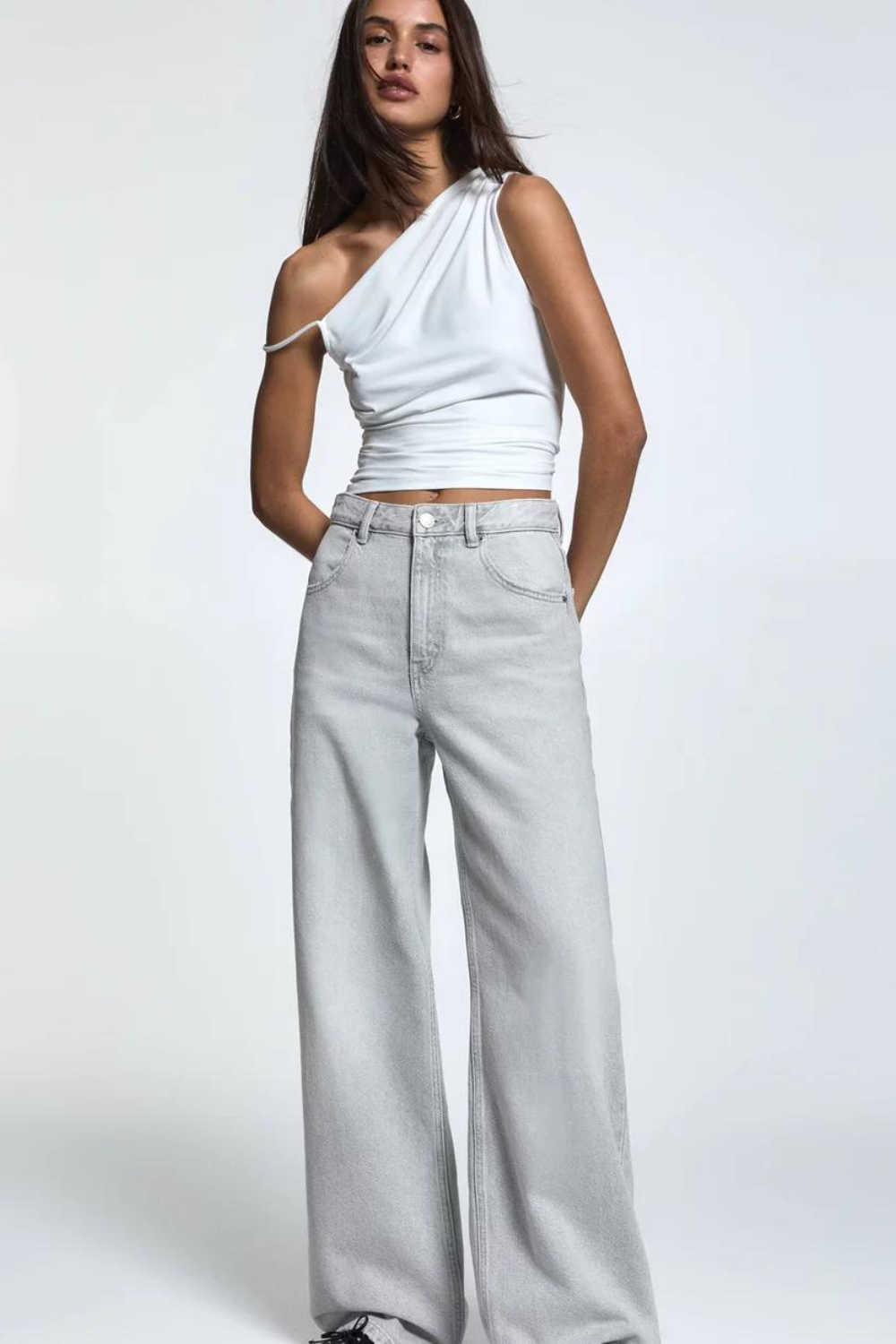 Person wearing a one-shoulder white top and grey wide-leg jeans, posing against a plain background.