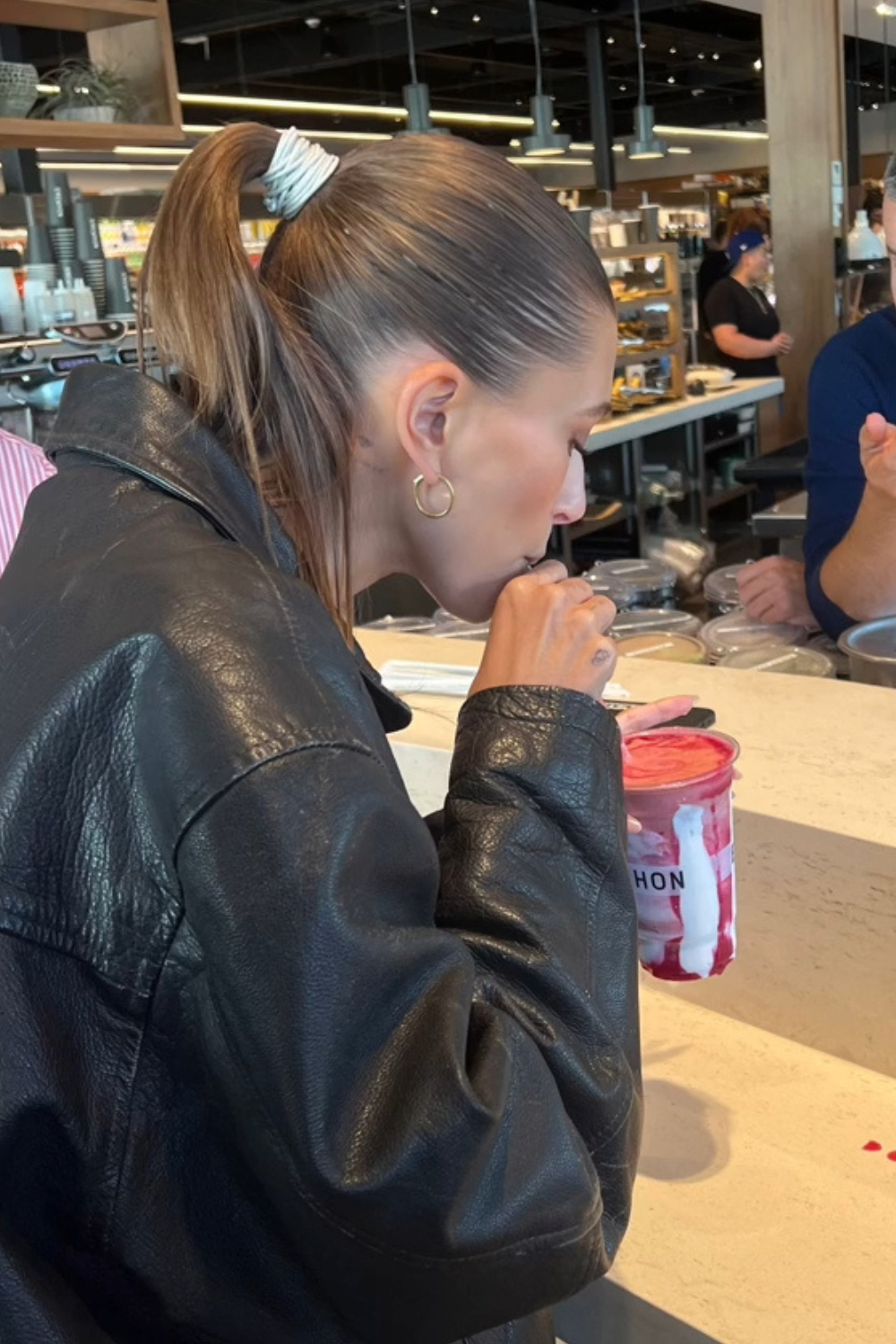 Hailey Bieber in a black jacket drinking a pink smoothie