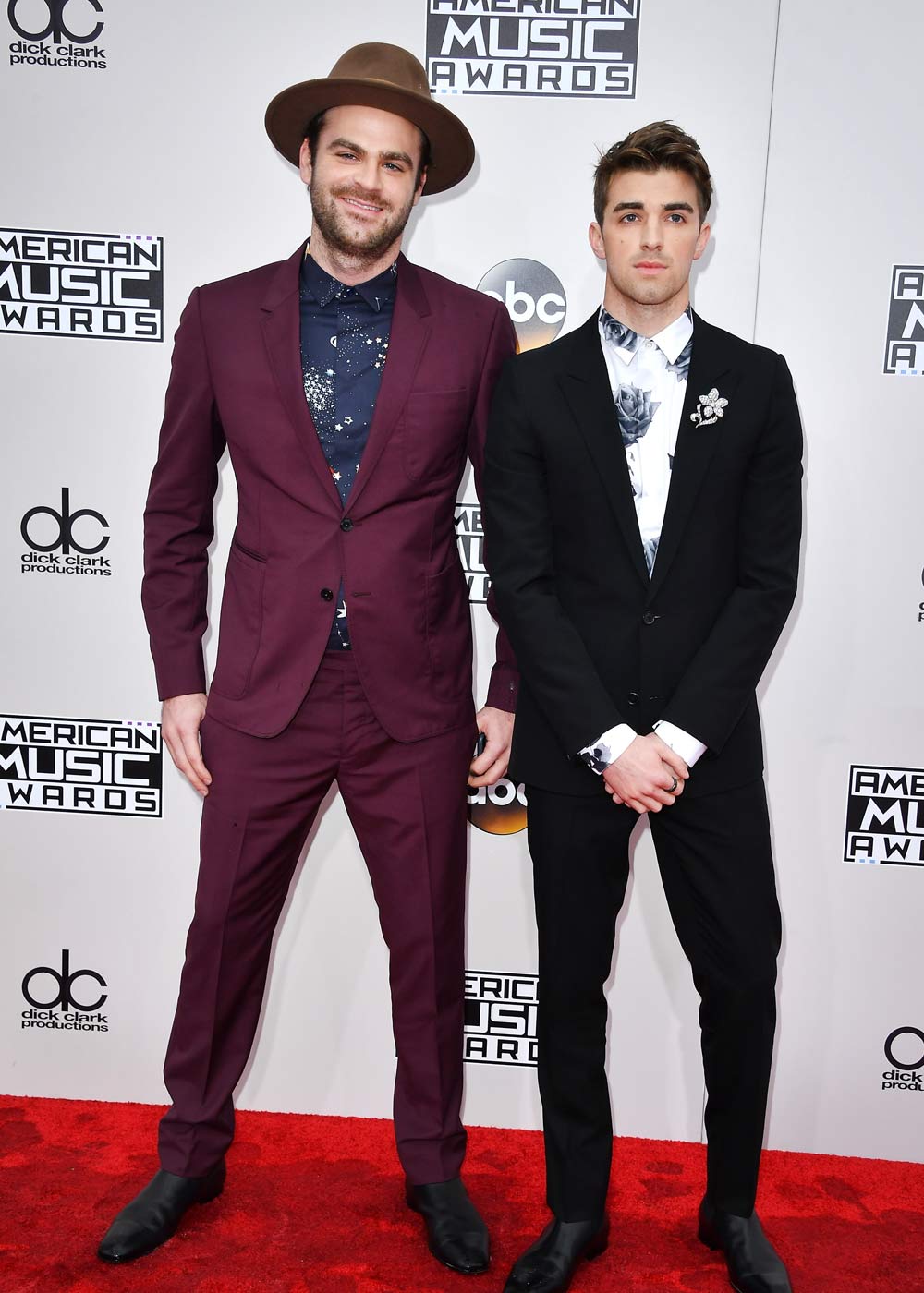 Two men pose on the American Music Awards red carpet; one in a burgundy suit and hat, the other in a black suit.