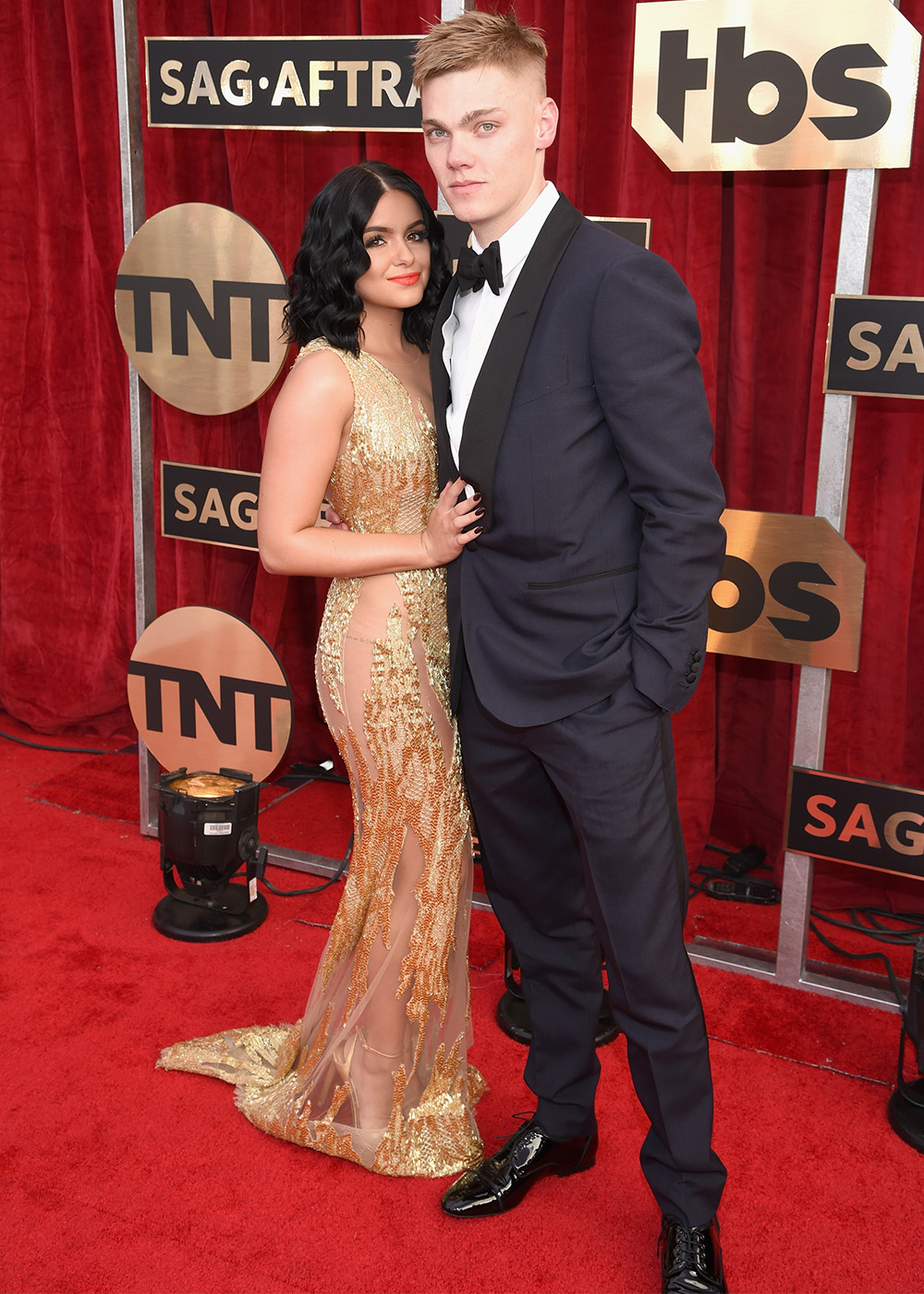 A woman in a gold dress and a man in a black suit pose on the red carpet at an awards event.