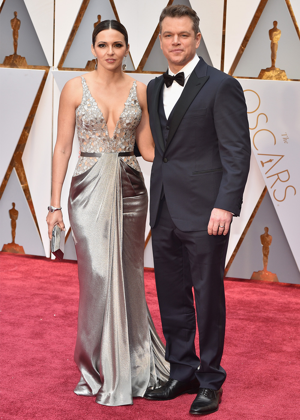 A woman in a silver gown and a man in a black tuxedo pose on the Oscars red carpet.